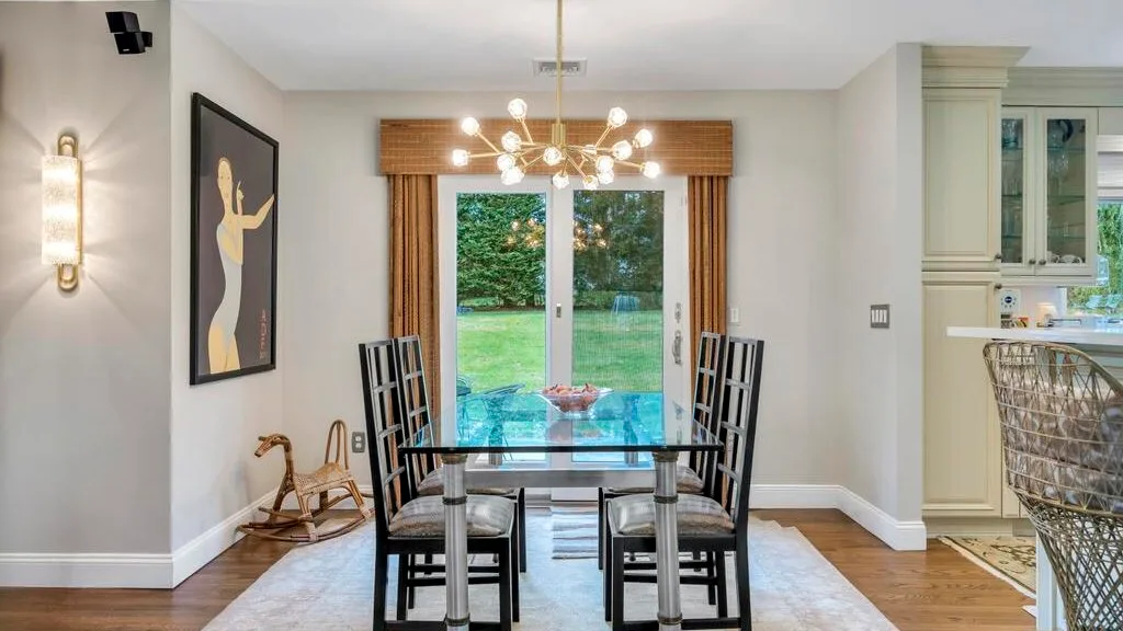 A dining area with a glass table and chairs beside large windows overlooking a garden.