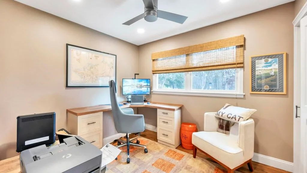 A home office with a desk, computer, and a cushioned seat beside the window.