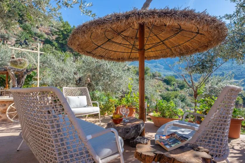An outdoor patio seating area sits beneath a large thatched parasol, surrounded by plants.
