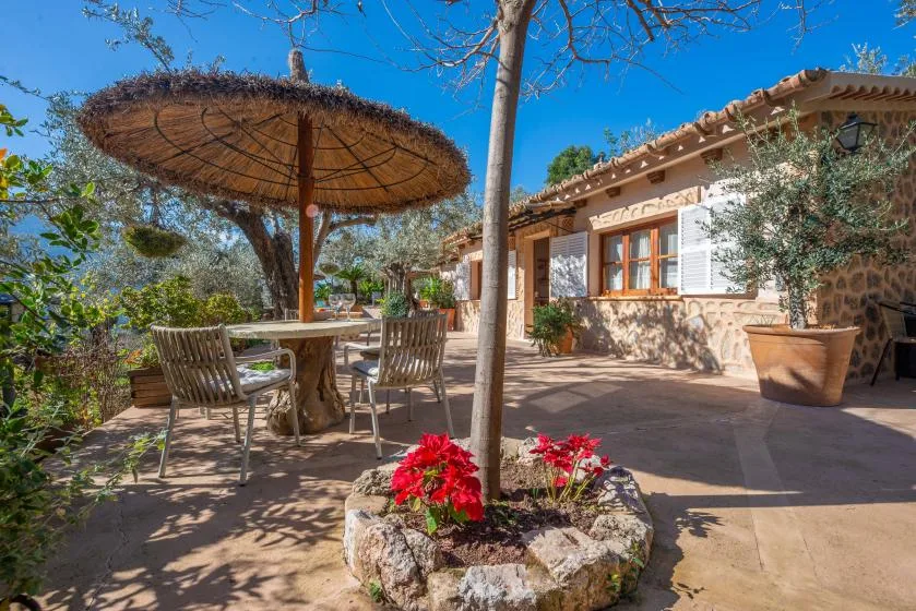 A sunny outdoor patio with seating beneath a thatched parasol beside a stone house.