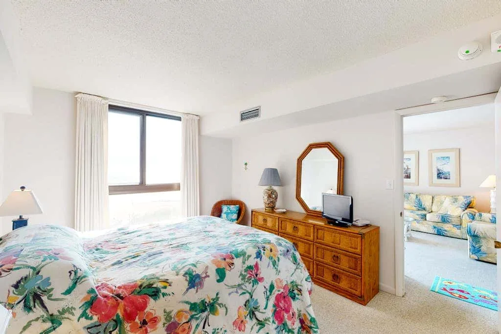A bedroom with a floral bedspread, dresser, and window overlooking a bright adjoining room.