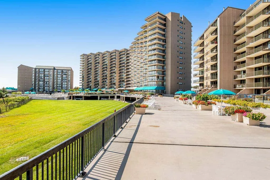 A paved outdoor terrace runs beside a grassy lawn and tall apartment buildings.