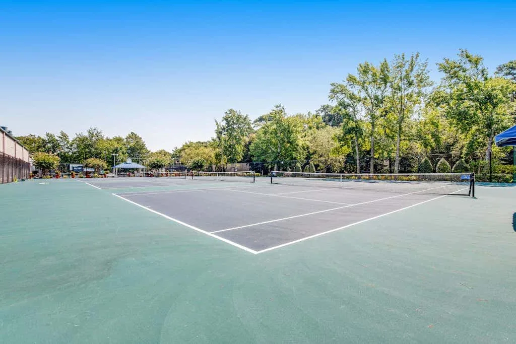An outdoor tennis court with white boundary lines and trees beyond the fence.