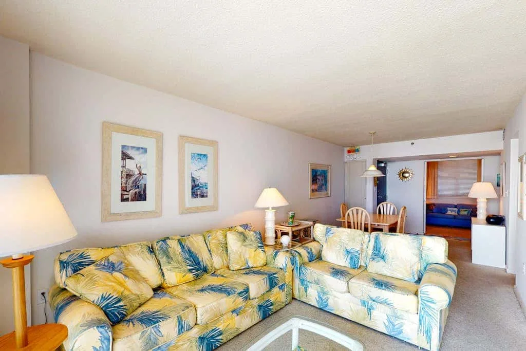 A living room with patterned seating, framed artwork, and a dining area beyond.