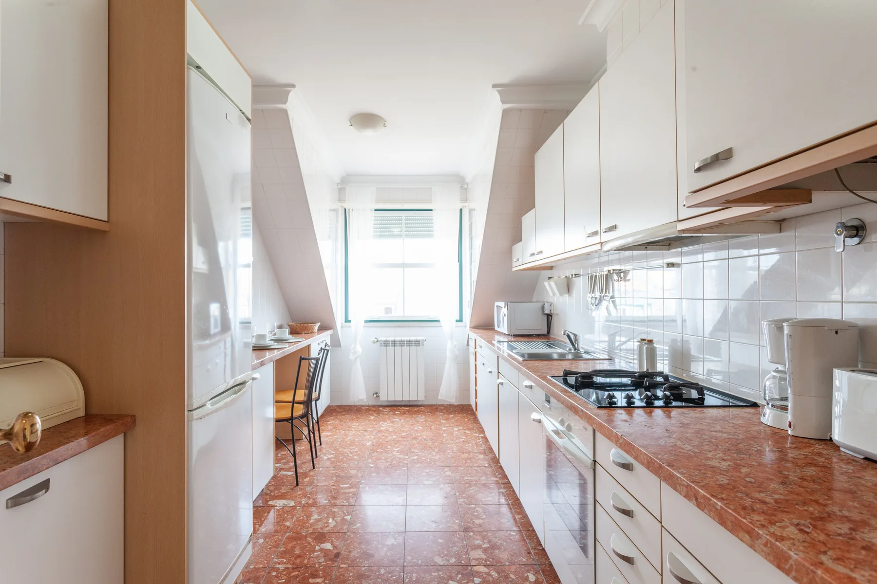 A narrow kitchen with white cabinets, countertop appliances, and a window at the far end.
