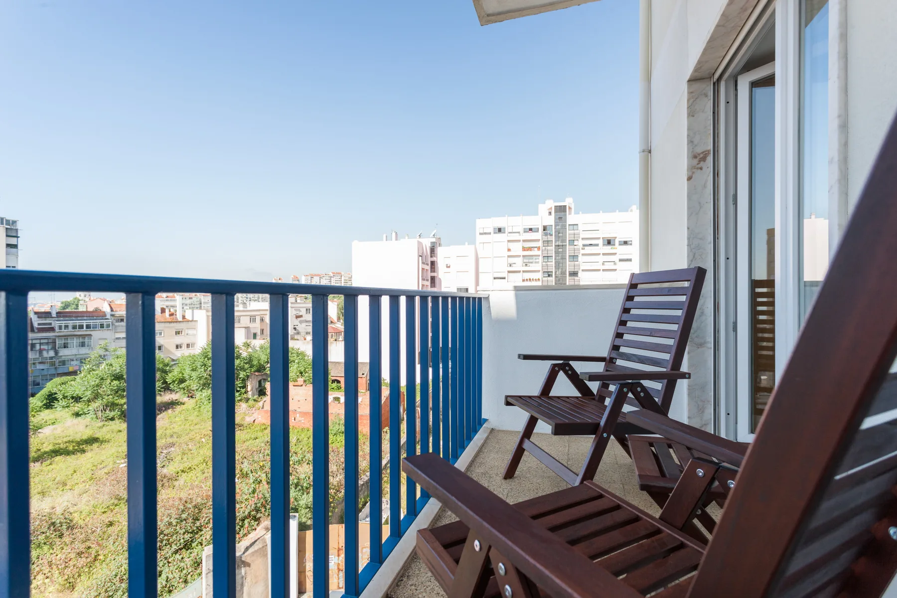 A balcony with wooden seating overlooks nearby buildings and greenery.