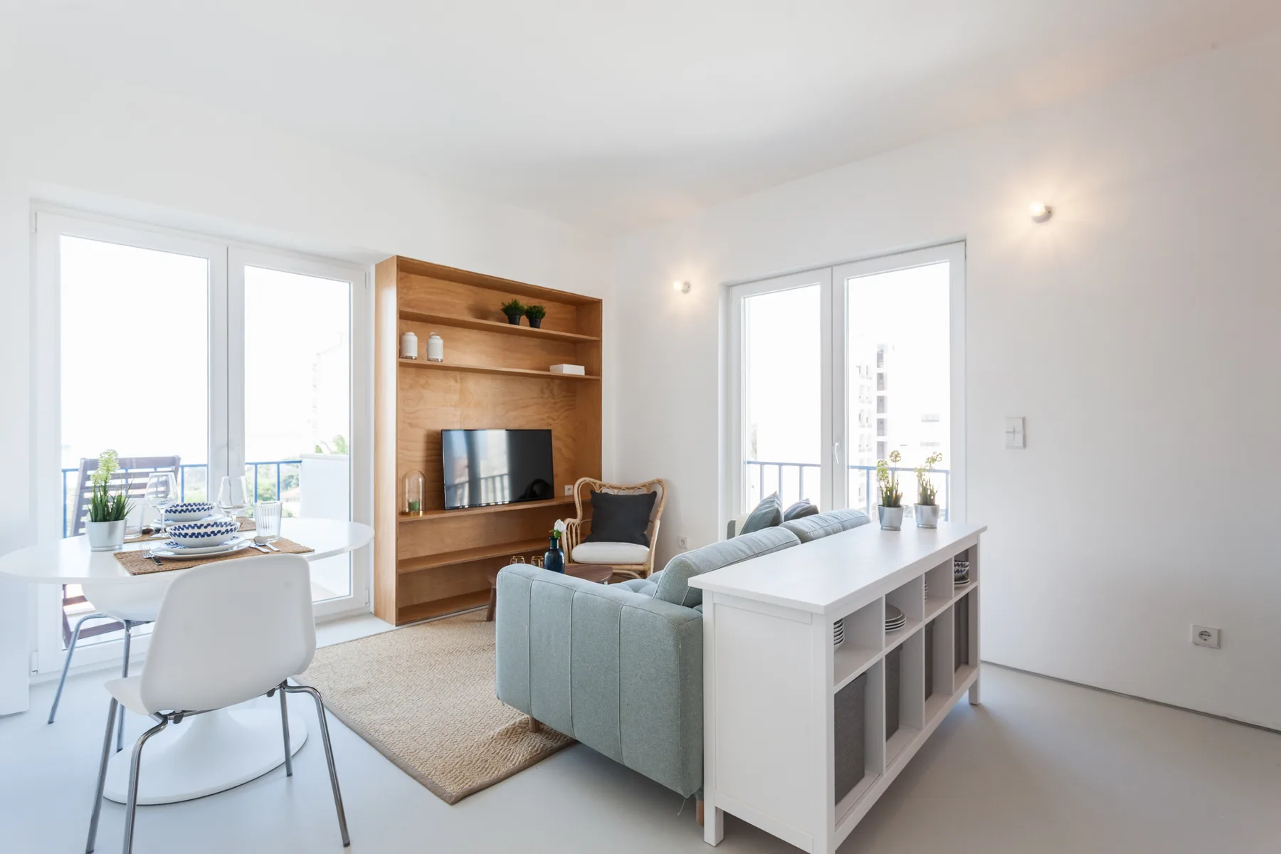 A bright living room has a dining table, seating, and a wooden shelving unit.