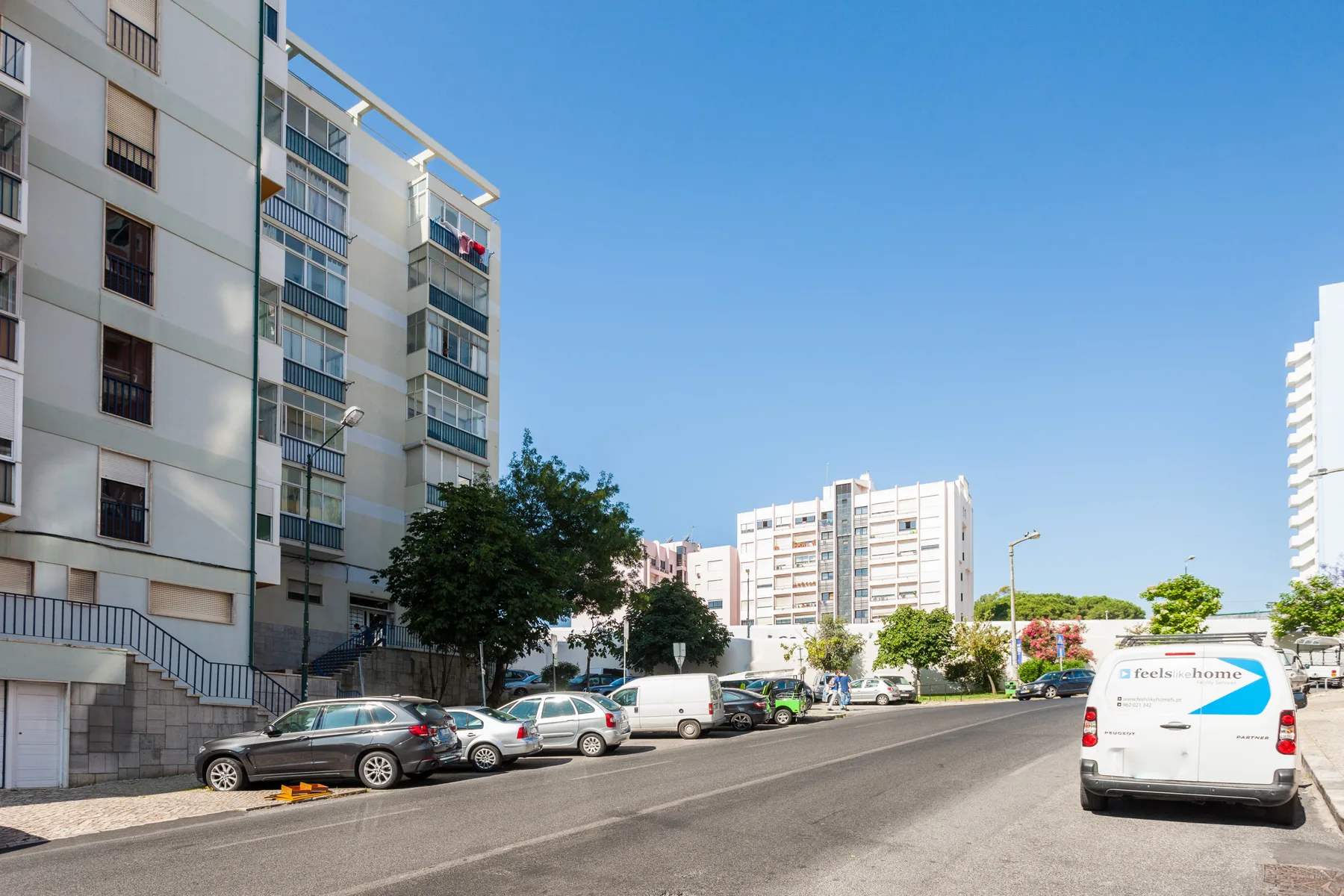 A city street with apartment buildings, parked cars, and a clear blue sky.