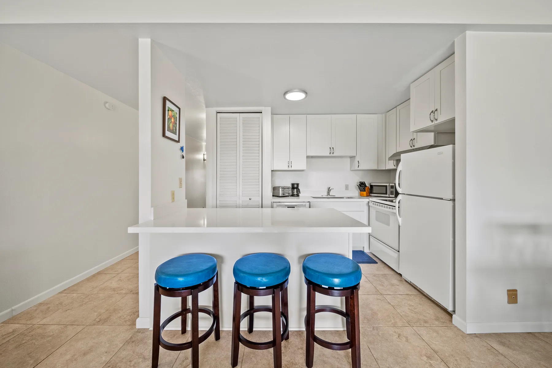 A bright kitchen with white cabinets, a tiled floor, and a breakfast counter with stools.