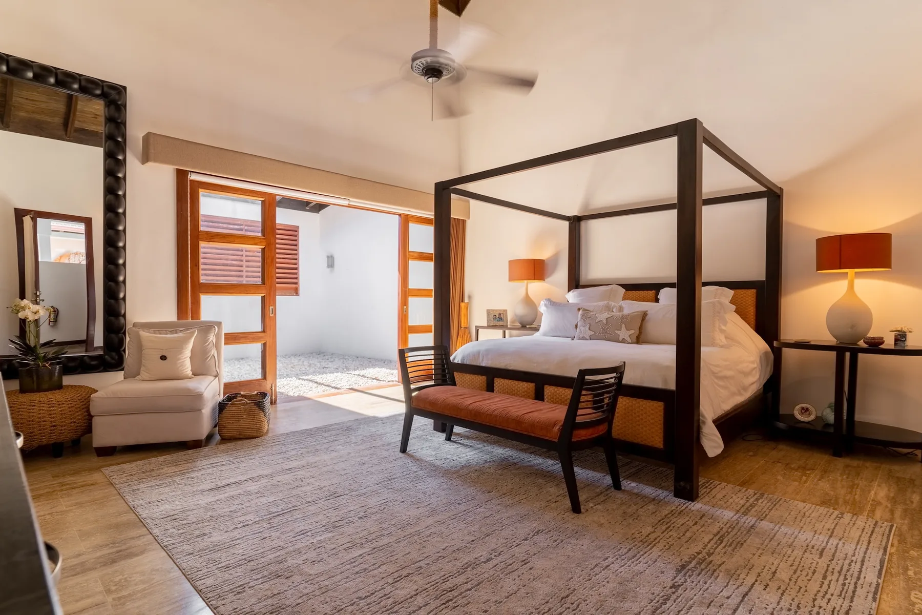 A bright bedroom with a four-poster bed and a seating area beside open doors.