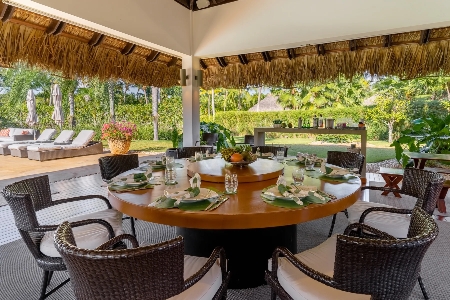 An outdoor dining area has a round table set beneath a thatched roof.