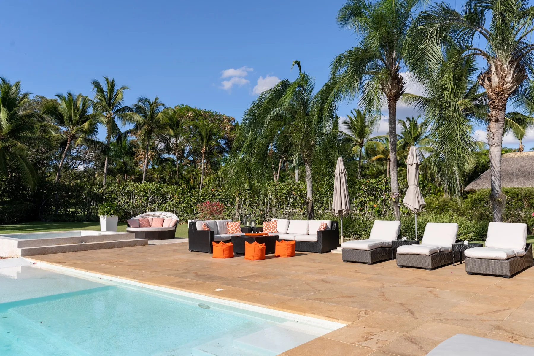 An outdoor pool beside a paved patio with lounge seating and tropical plants.