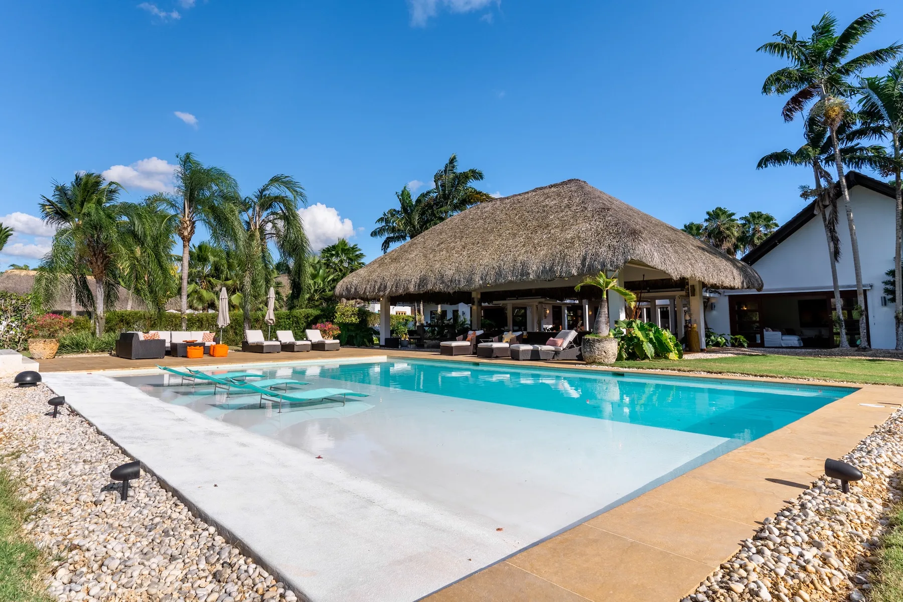 A swimming pool beside an open-sided thatched-roof pavilion and palm trees.