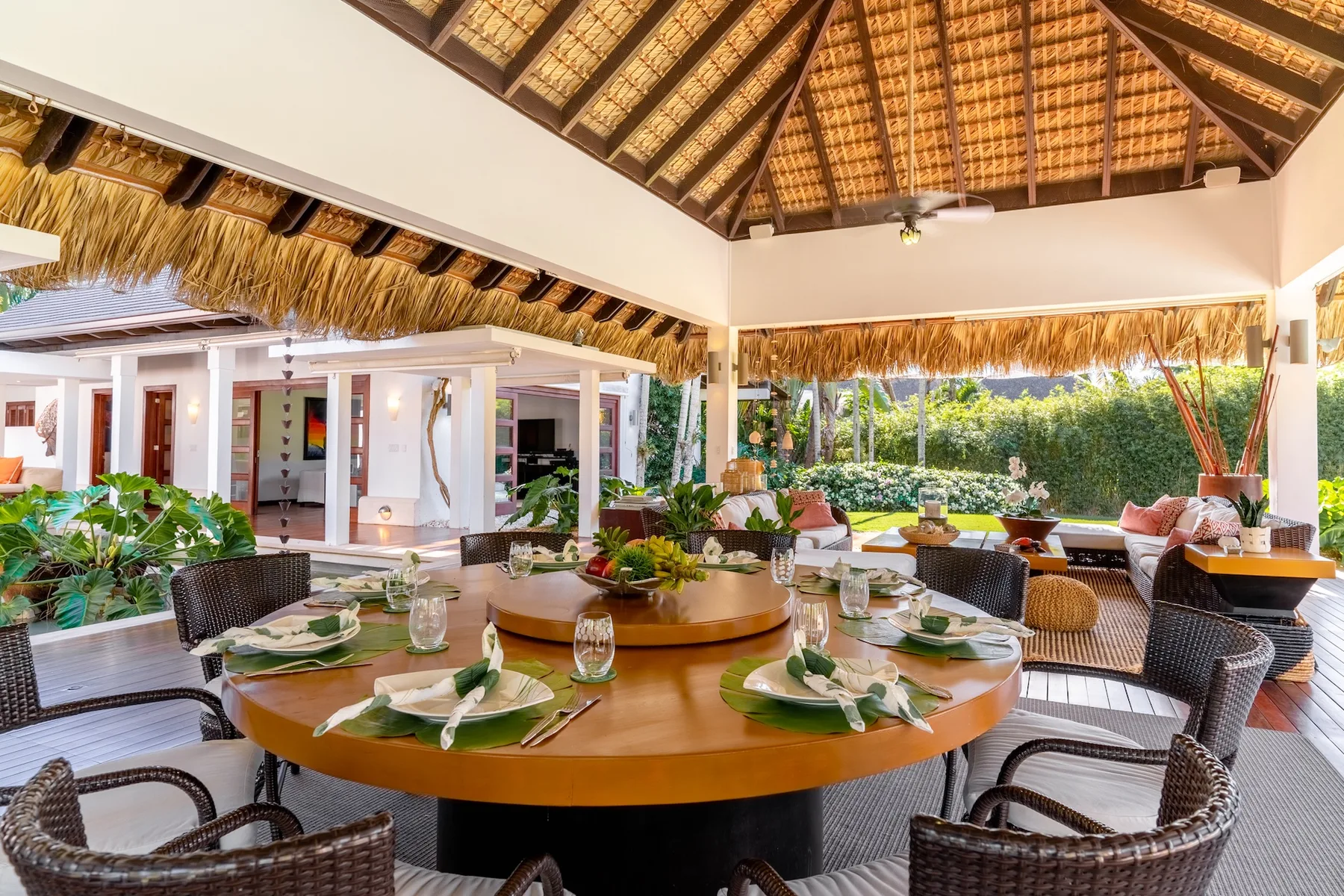 An open-air dining area has a round table set with plates and greenery.