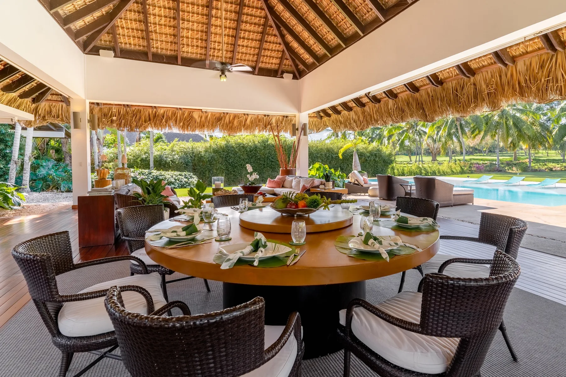 An open-air dining area with a round table set beside a garden and pool.