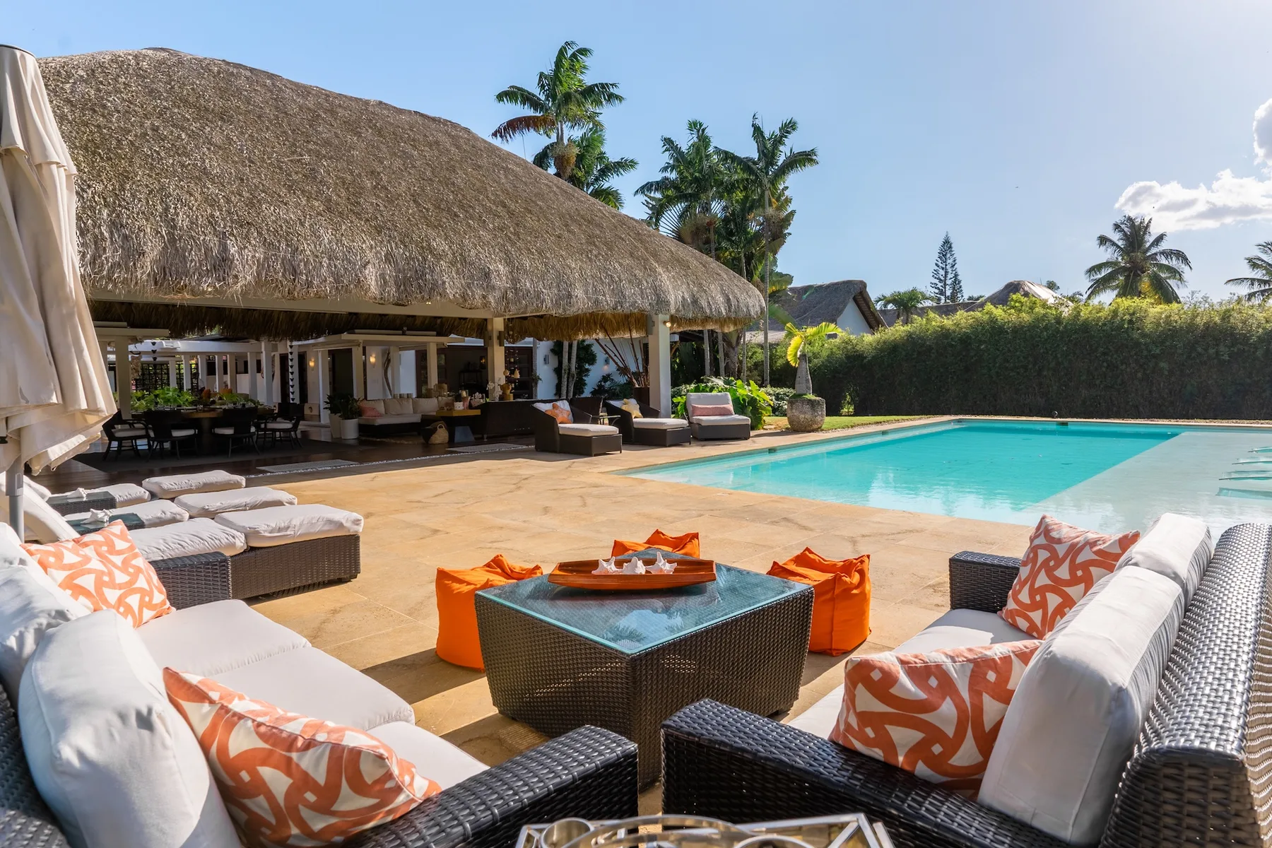 A poolside patio with outdoor seating, a thatched-roof pavilion, and palm trees.