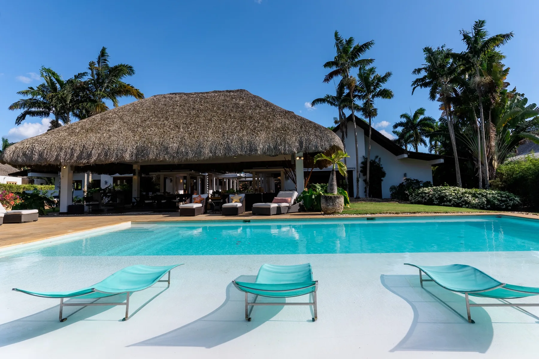 A resort pool with lounge seating in front of a thatched-roof building.