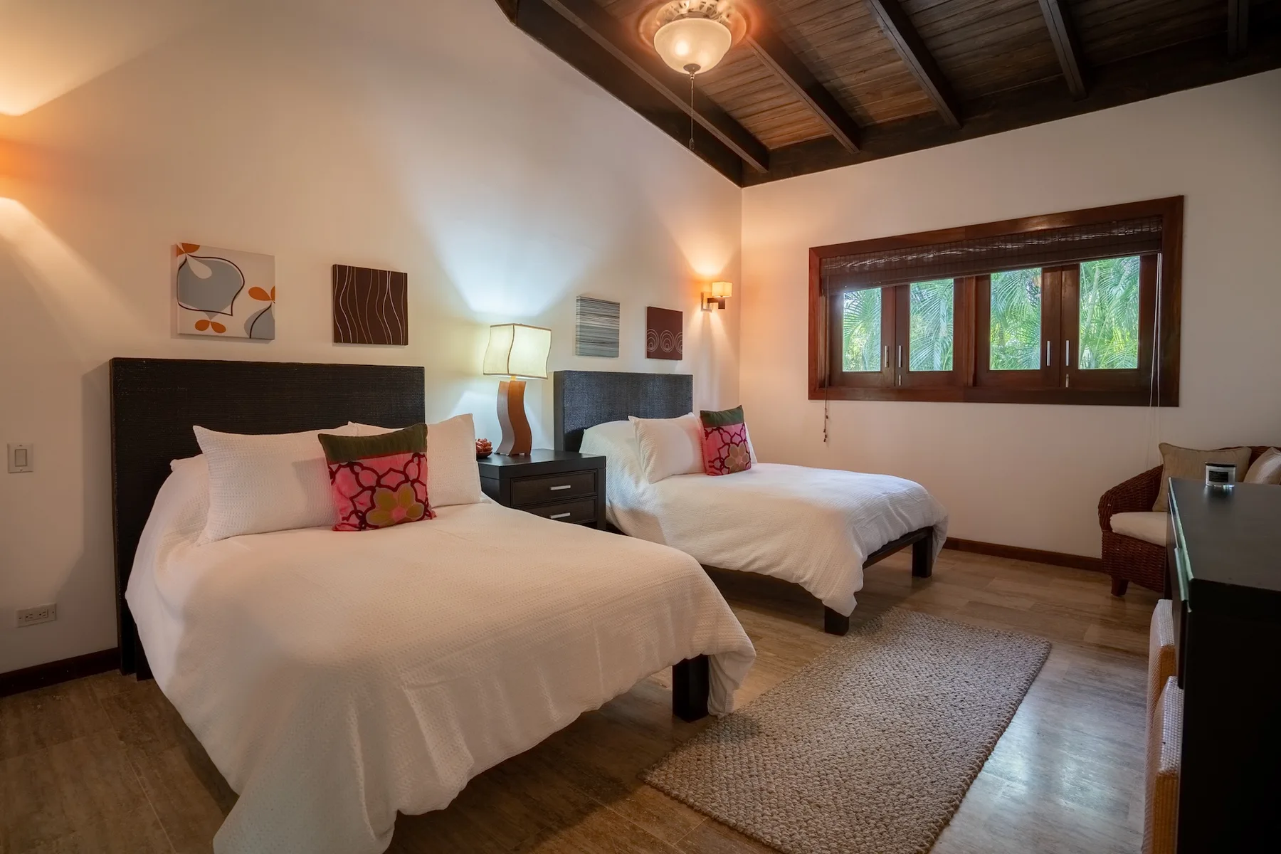 A bedroom with two beds, bedside tables, and a window overlooking greenery.