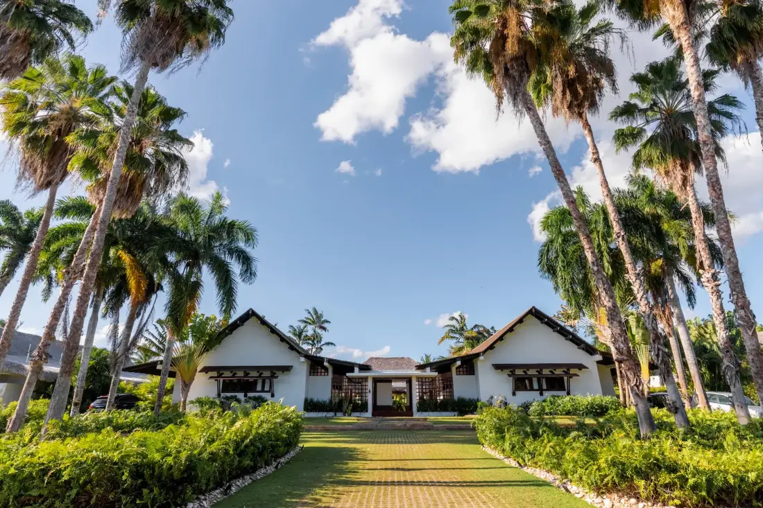 A low white building stands beyond a landscaped lawn, framed by palm trees.