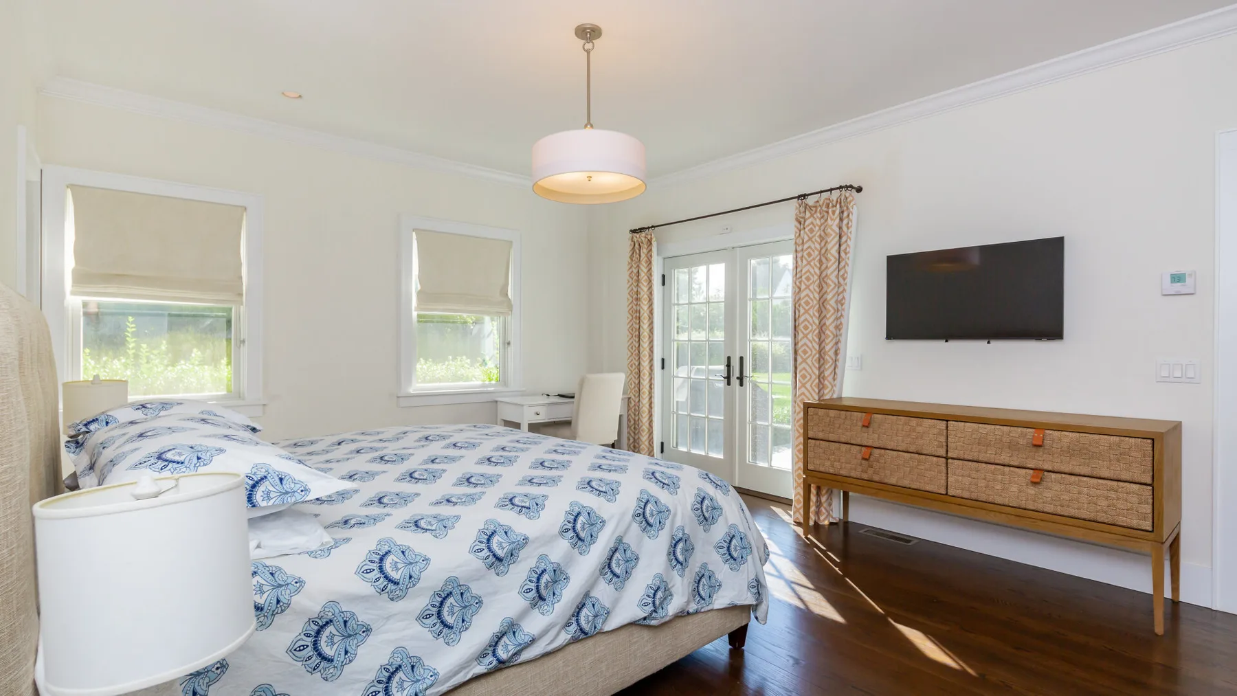 A bright bedroom with a patterned bedspread, windows, and a wall-mounted television.