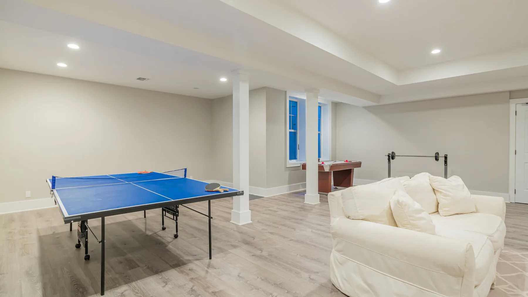 A basement recreation room with a blue ping-pong table and white seating.