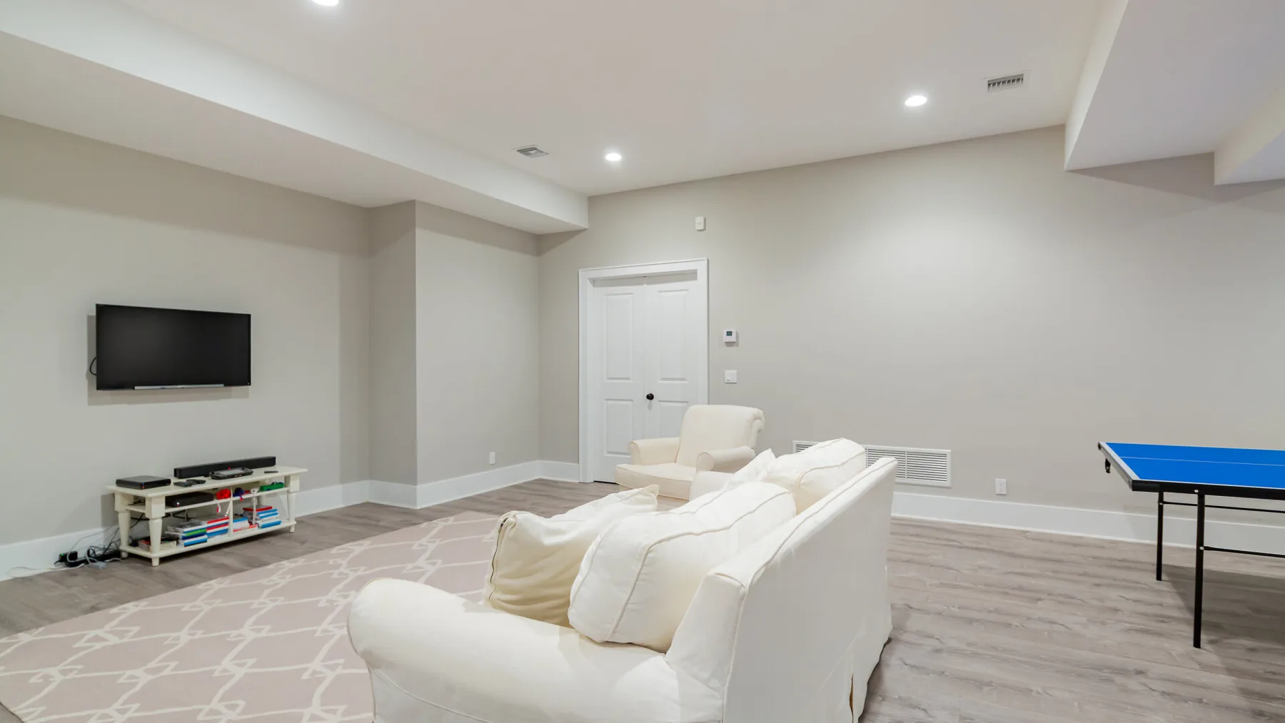 A bright living room with white seating, a wall-mounted television, and a blue table tennis table.