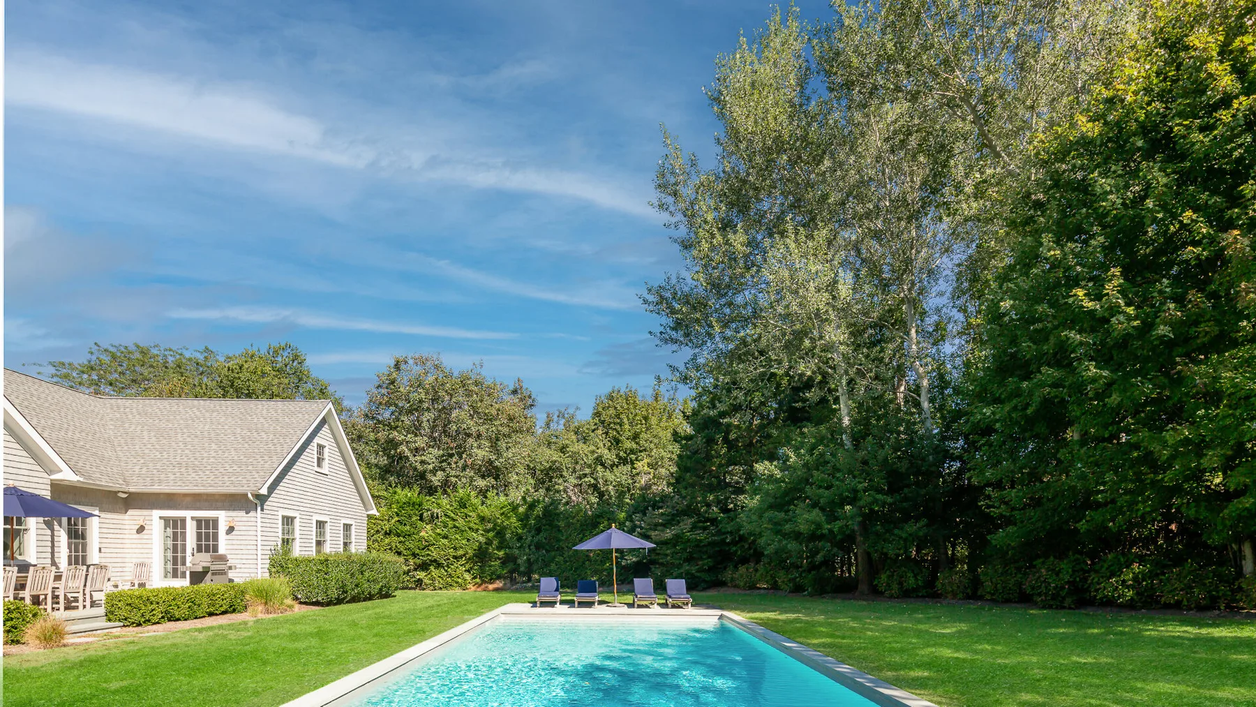 A swimming pool beside a light-colored house, with trees and lawn around it.