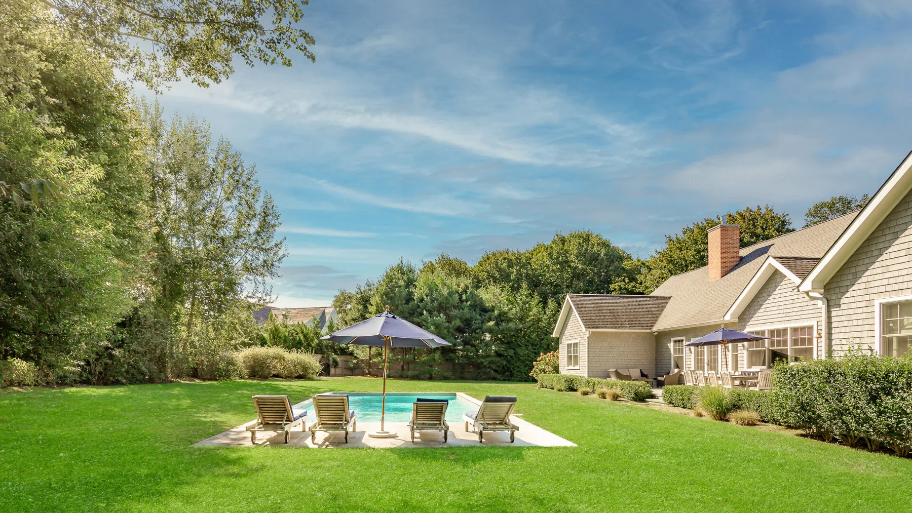 A landscaped backyard features a swimming pool, patio seating, and a large house.