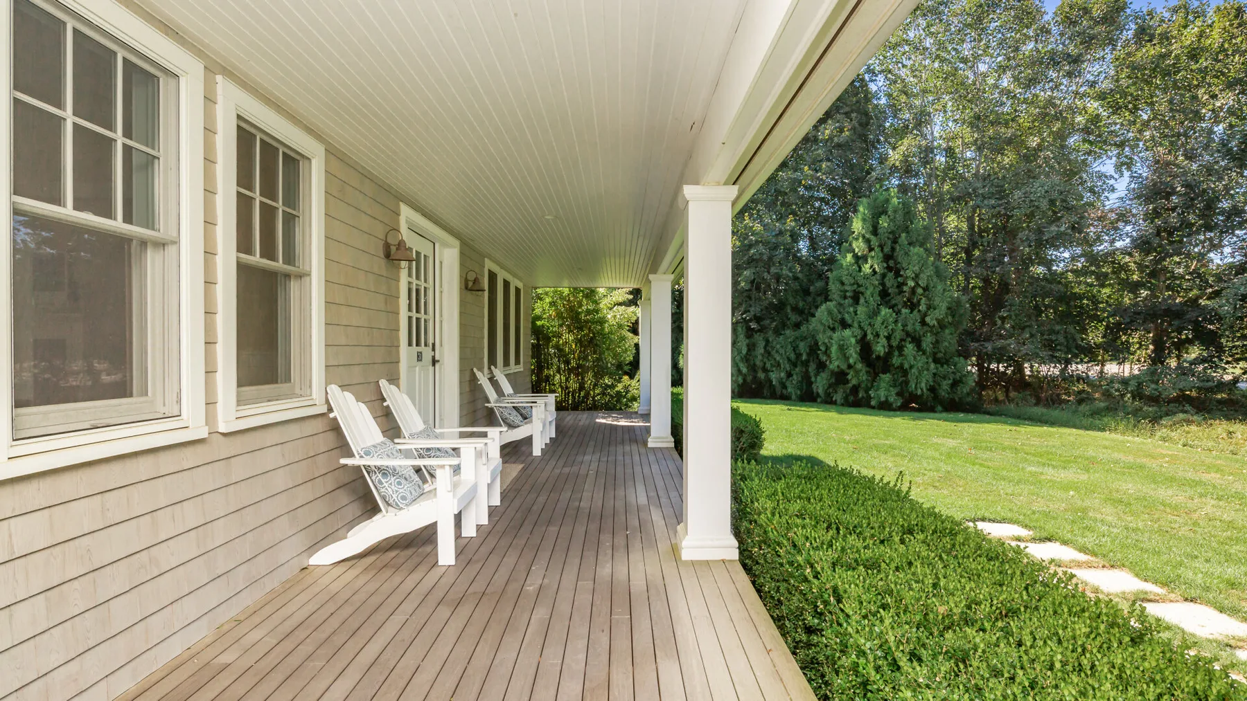 A covered porch with white seating overlooks a green lawn and trees.