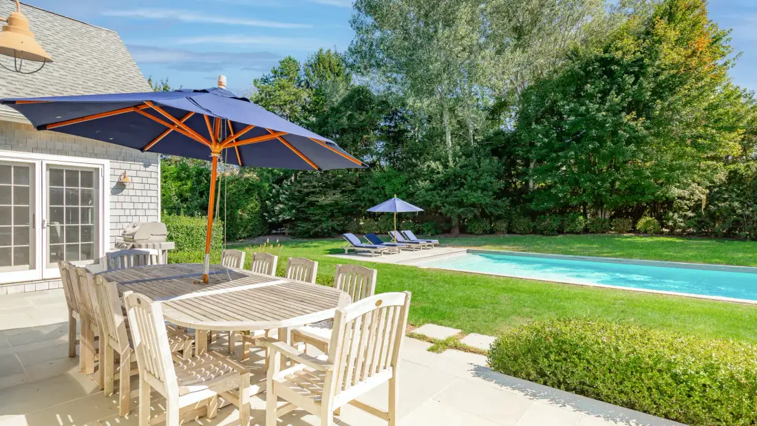 An outdoor dining area with a large umbrella overlooks a swimming pool.
