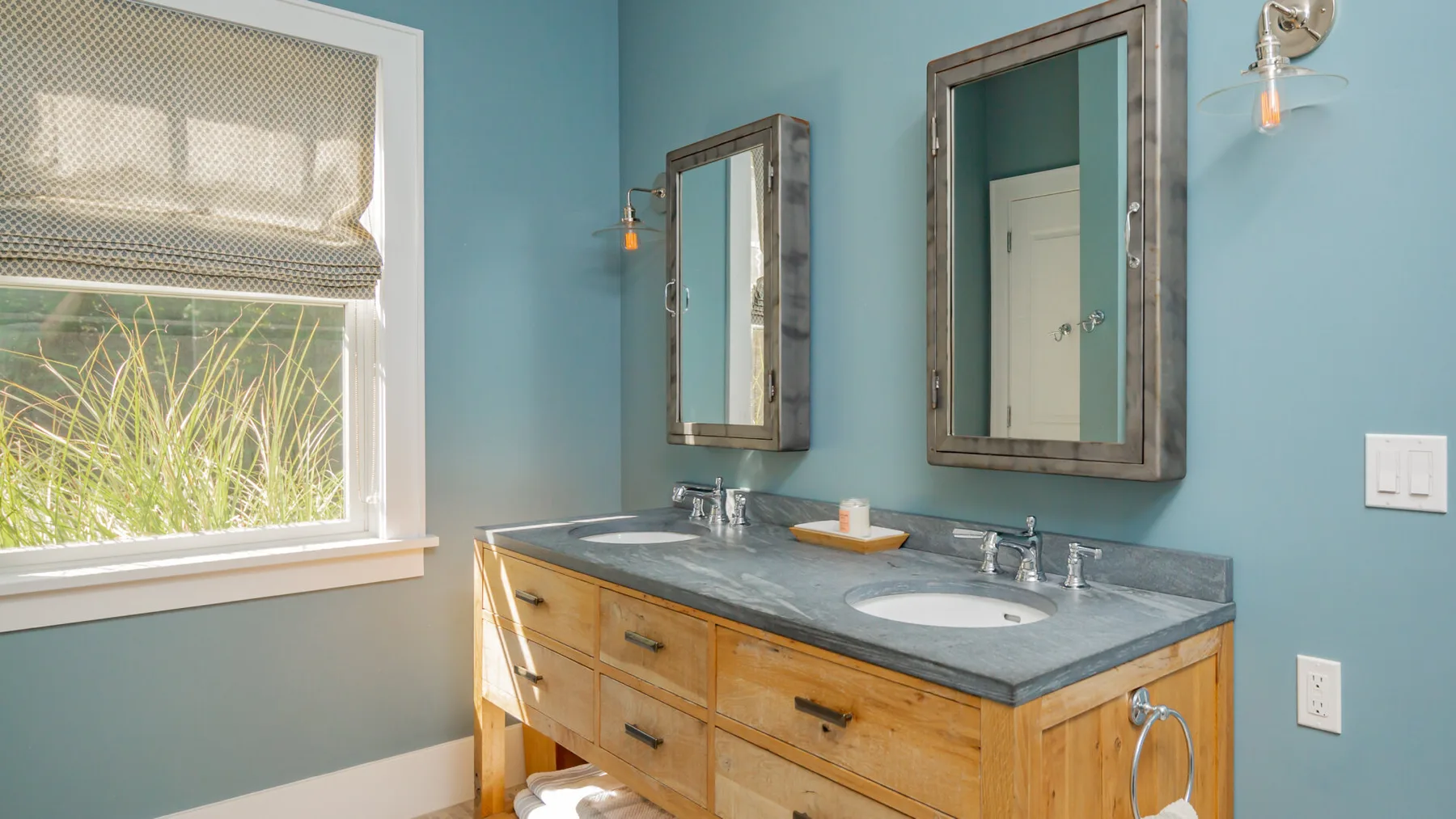 A bathroom vanity with two sinks sits beneath mirrors beside a window.