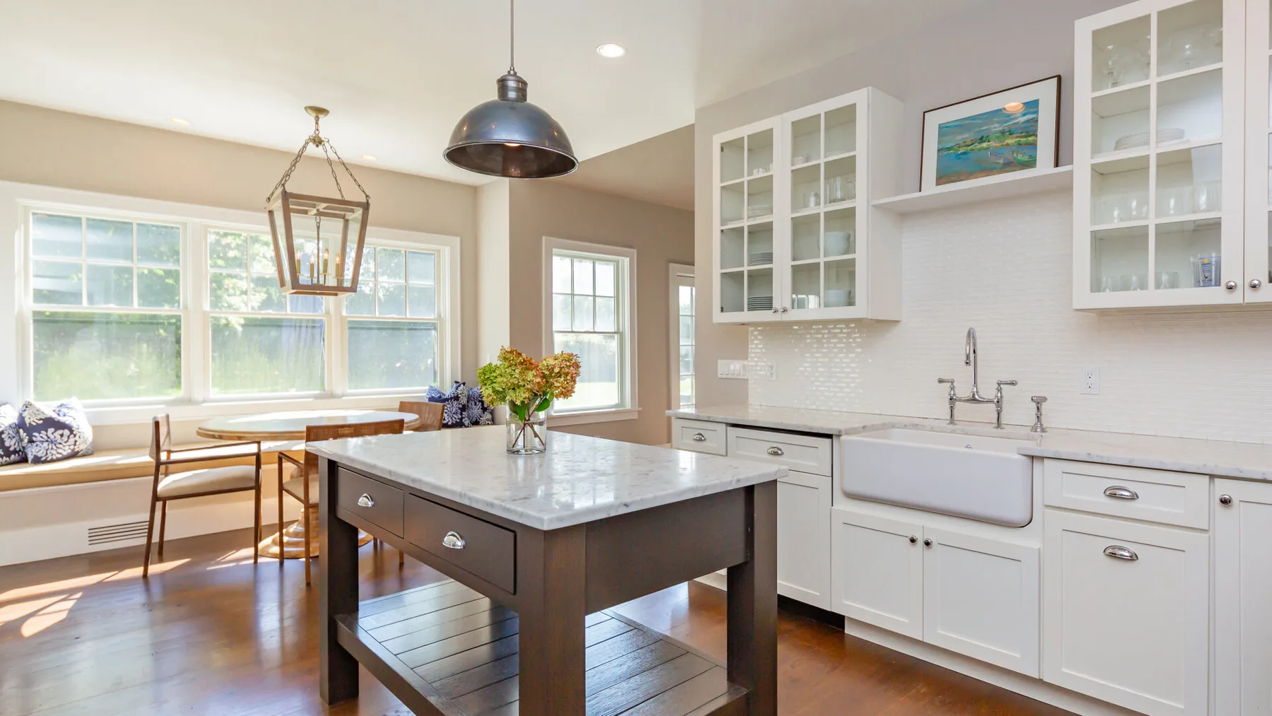 A bright kitchen with a central island, white cabinetry, and windows overlooking a dining nook.