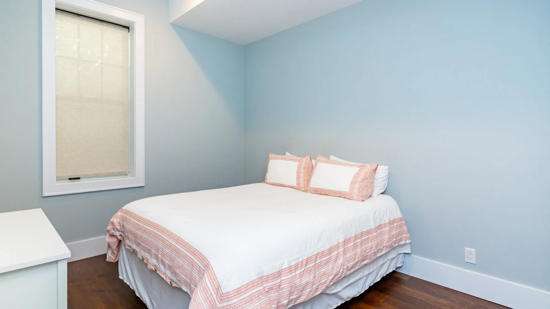 A bedroom with a bed dressed in white and pink bedding beside a window.