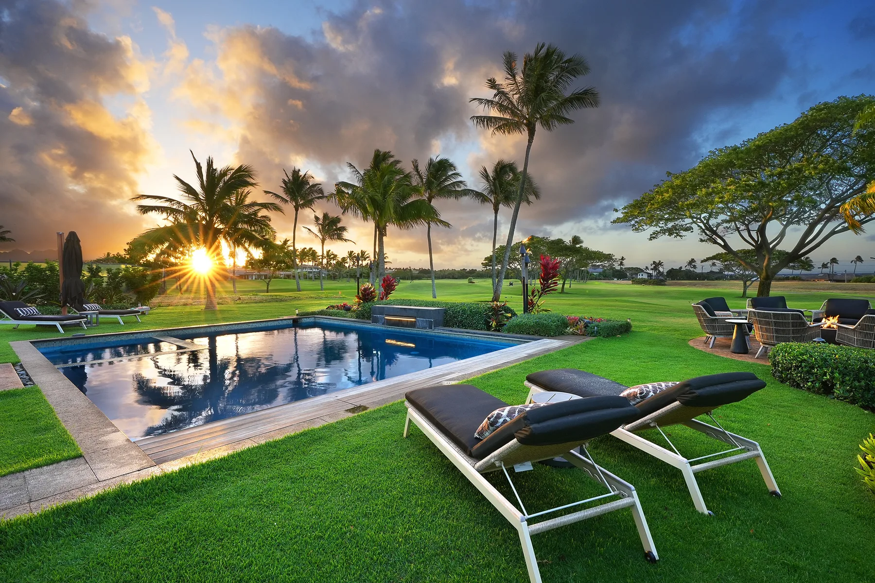 An outdoor swimming pool and lounge seating overlook a tropical lawn at sunset.