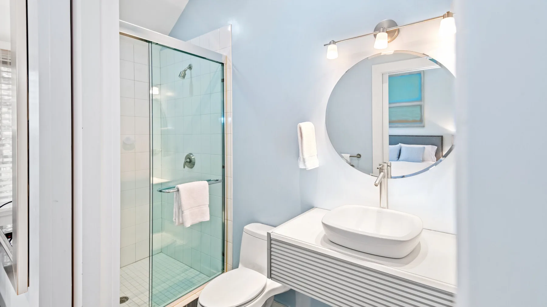 Un baño luminoso con ducha de vidrio, inodoro y tocador con espejo redondo.