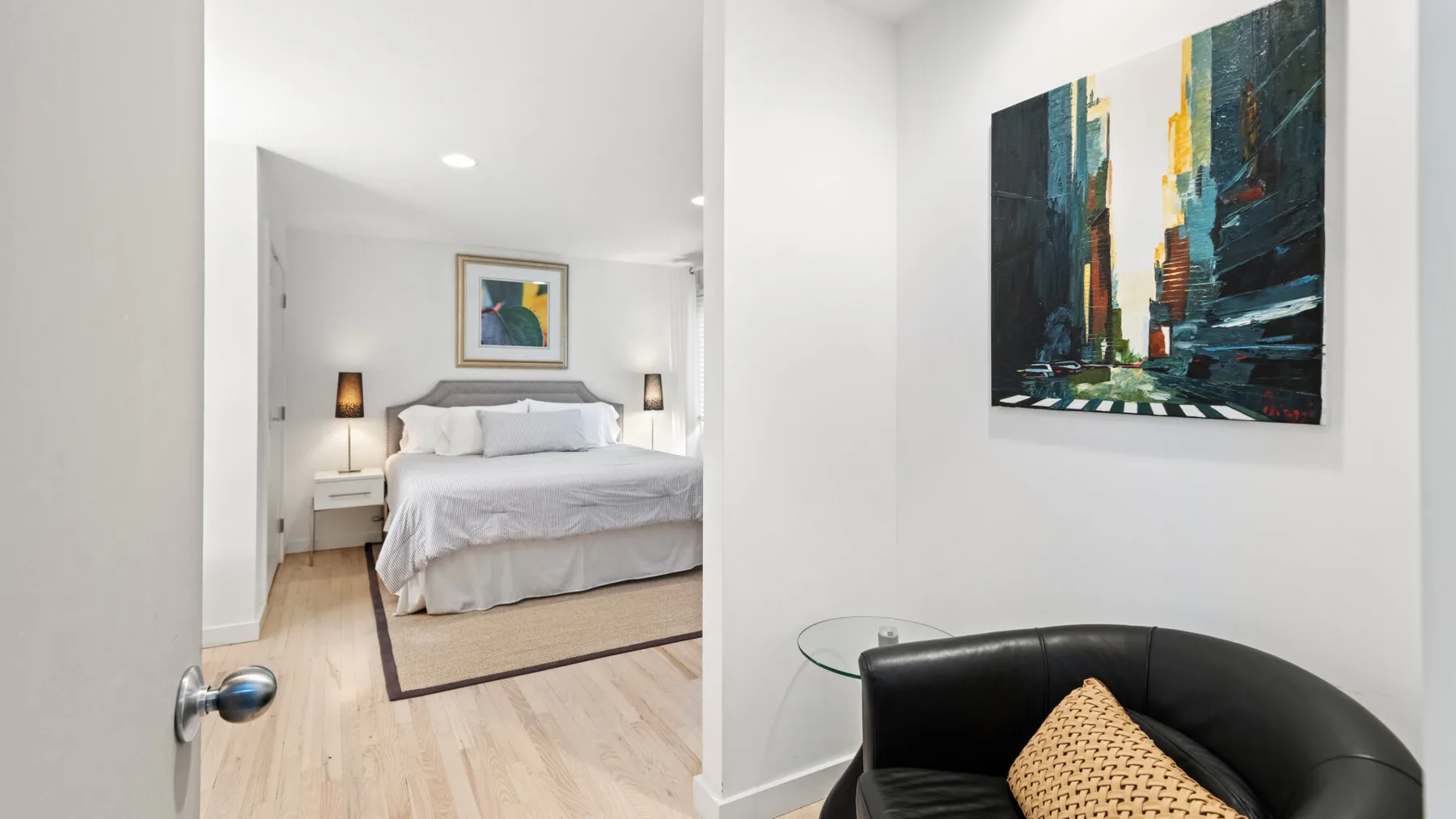 Un dormitorio con una cama tendida y un sillón negro junto a un cuadro colorido.