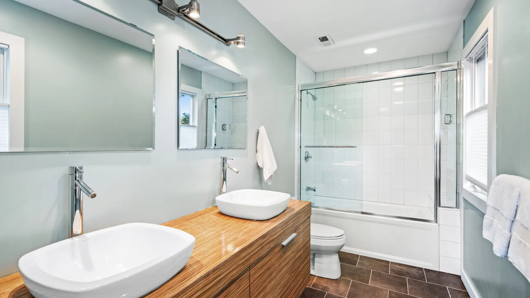 Un baño luminoso con tocador de madera, lavabos sobre encimera y bañera con ducha y mampara de vidrio.