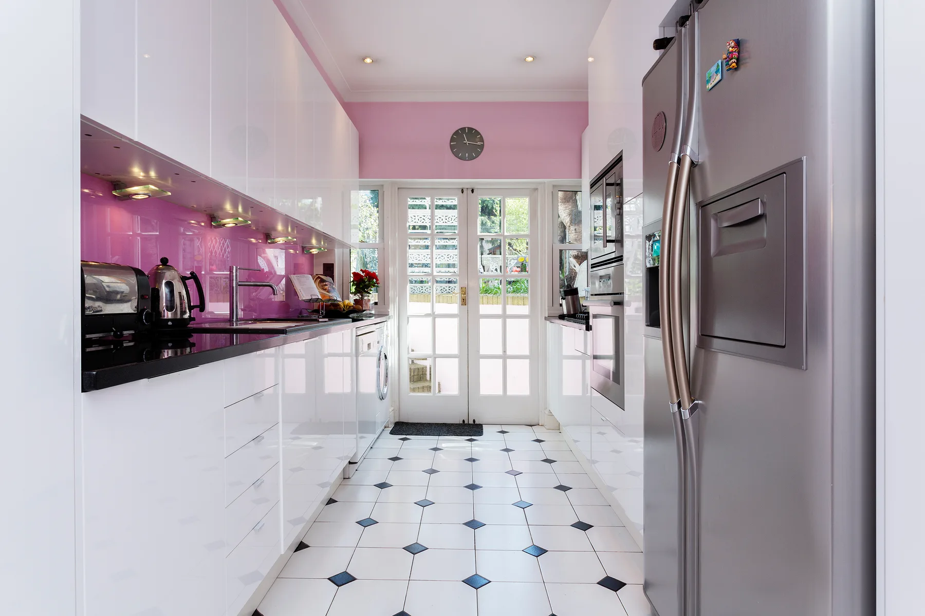 A bright galley kitchen has white cabinets, a pink backsplash, and glass doors at the far end.