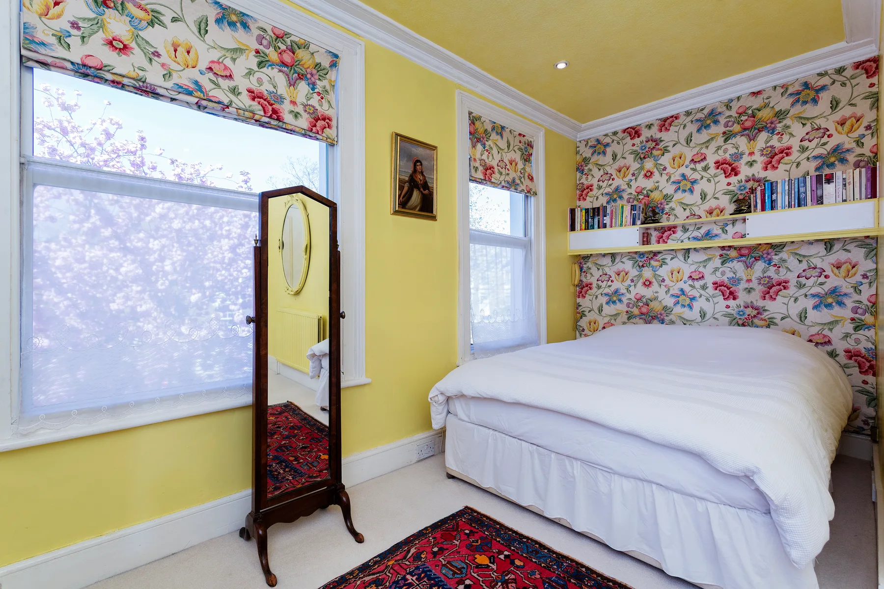 A bedroom with a white bed, floral wallpaper, and large windows.