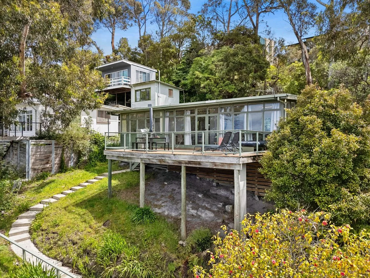 A modern house with a glass-walled patio sits among trees and garden plants.