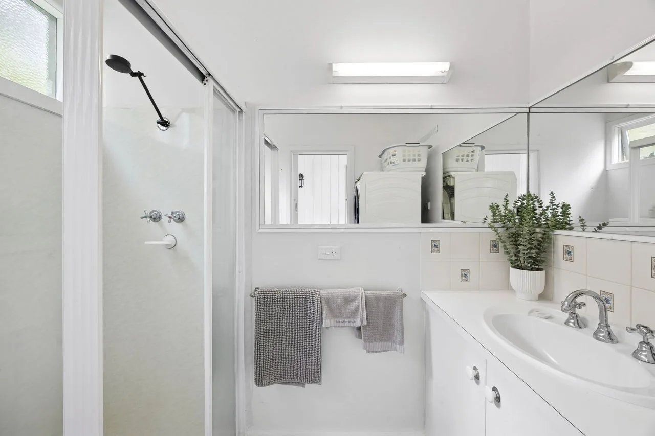 Un baño luminoso con ducha de vidrio, un espejo grande y un tocador blanco.