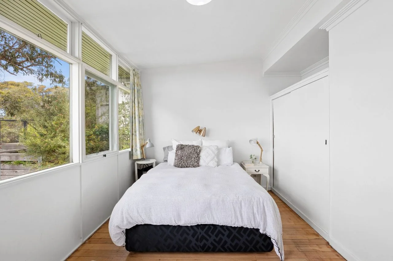 Un dormitorio luminoso con una cama junto a grandes ventanas que dan a los árboles.