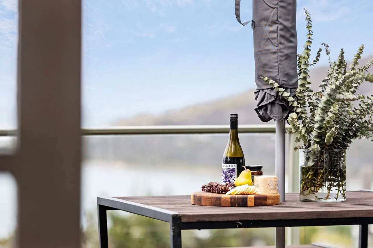 Una mesa de balcón tiene una botella, fruta y flores junto a una vista abierta.