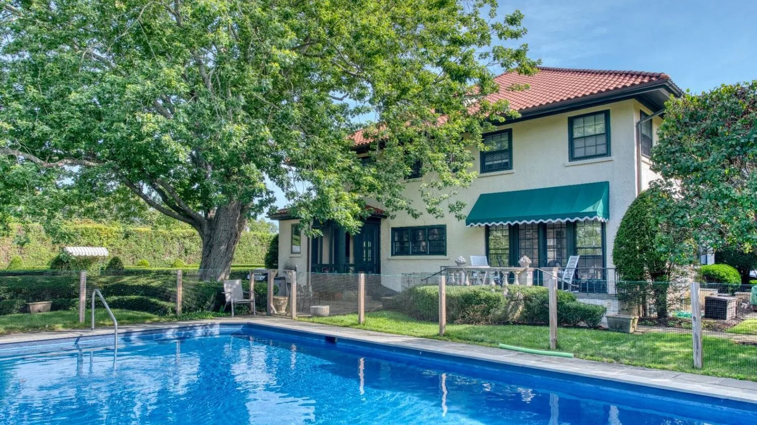 Una piscina azul está junto a una casa de dos pisos y un jardín frondoso.