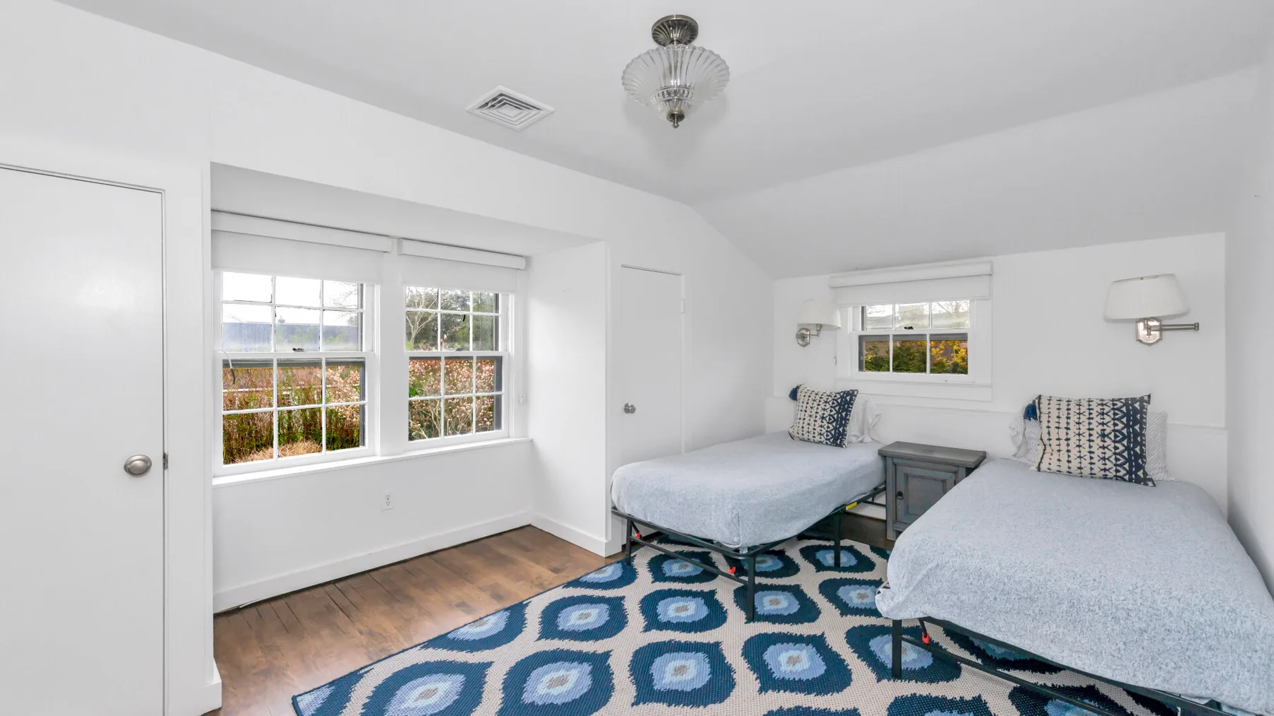 Un dormitorio luminoso con dos camas, ventanas grandes y una alfombra azul estampada.