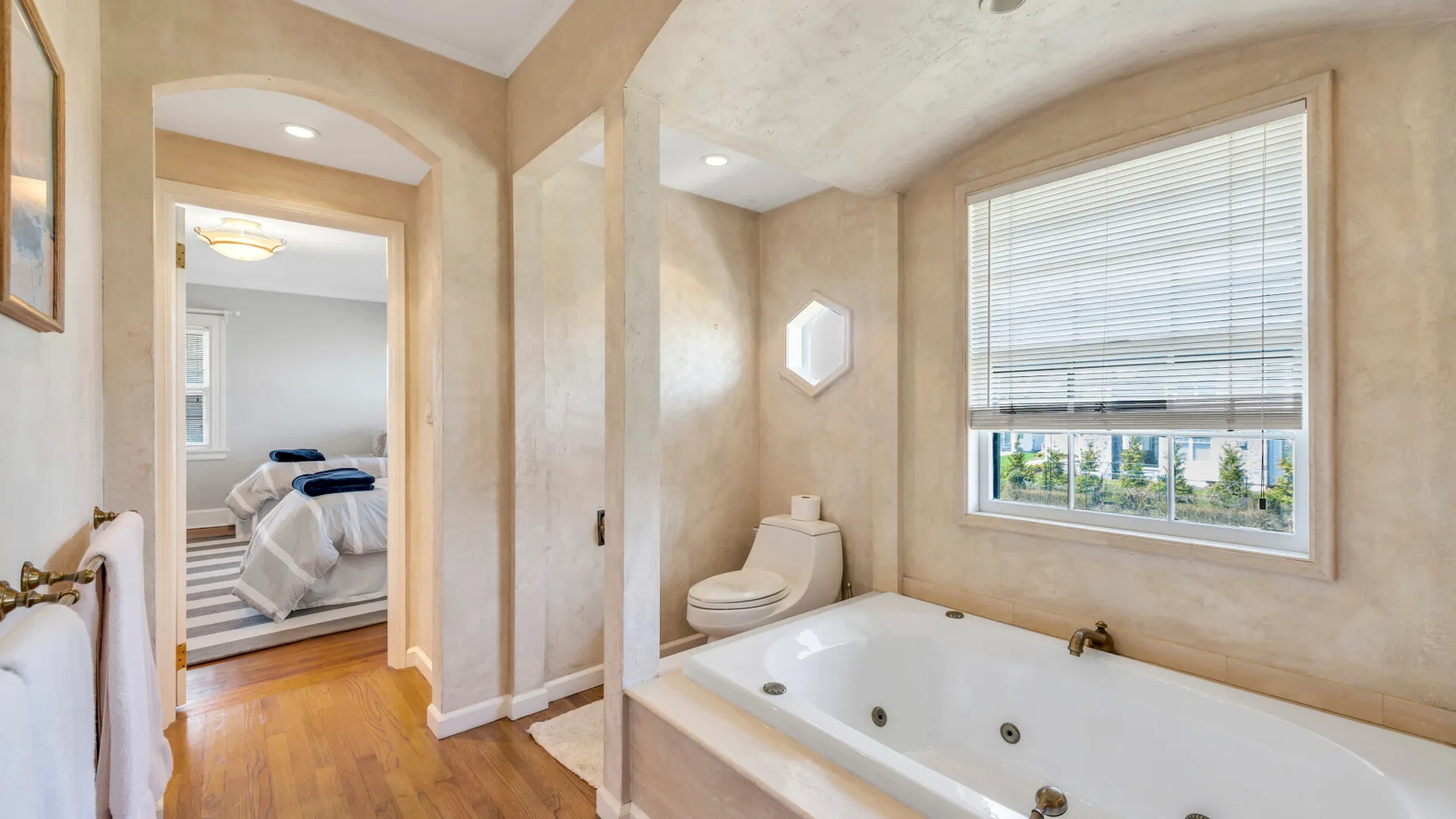 Un baño con bañera junto a una ventana grande y una puerta abierta hacia un dormitorio.