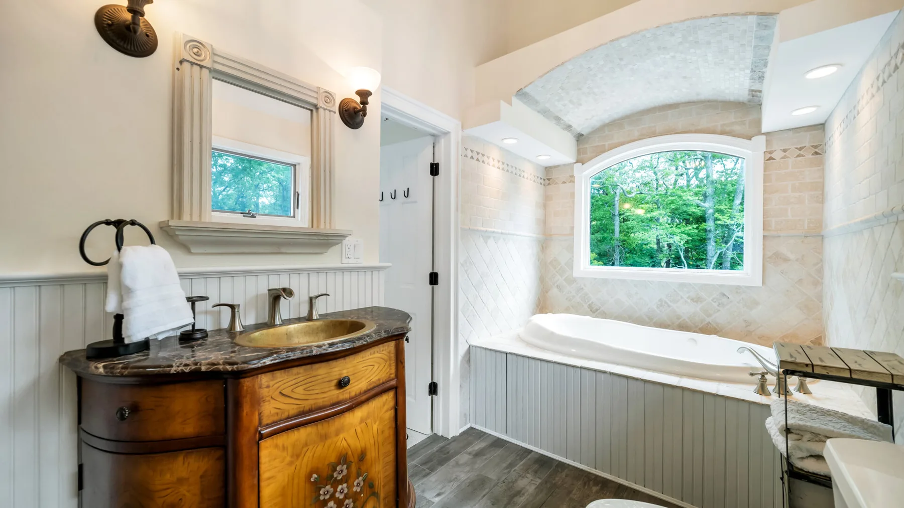 Un baño luminoso cuenta con un tocador de madera y una bañera bajo una ventana grande.