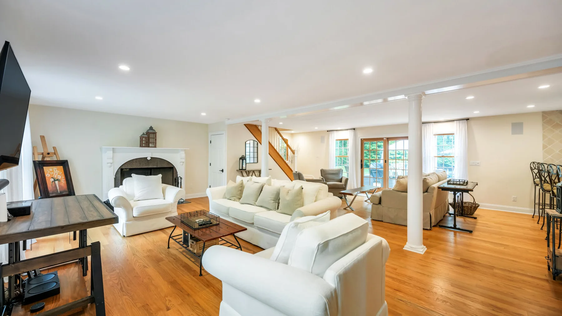 A bright living room with white seating, wooden floors, and an open staircase.
