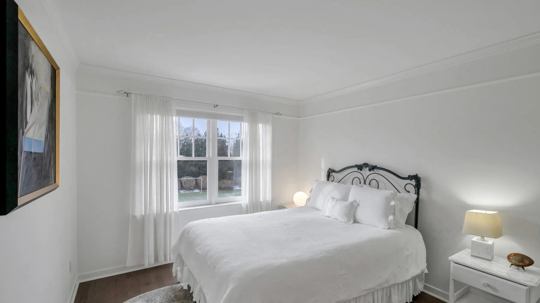 Un dormitorio luminoso con una cama blanca junto a una ventana con cortinas translúcidas.