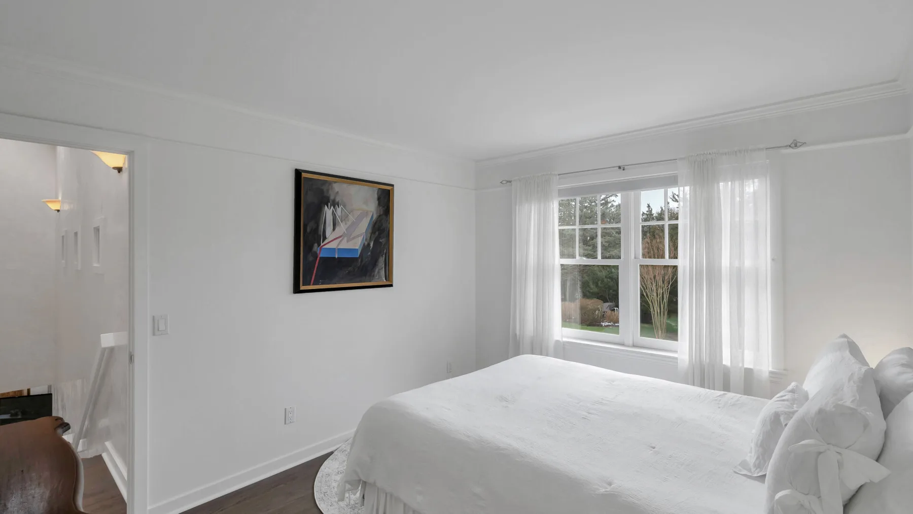 Un dormitorio luminoso con una cama blanca junto a una ventana con cortinas transparentes.